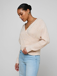 A woman wears the Antonella Crossover Sweater in light pink, featuring small textured dots, paired with light blue jeans. She stands against a plain off-white background, looking down and slightly to the side.