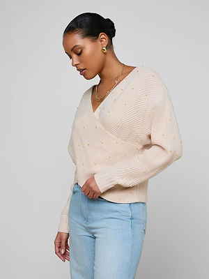 A woman in the Antonella Crossover Sweater, a light beige knit with pearl accents and subtle embellishments, pairs it with light blue jeans as she stands against a plain light gray background, looking down with one hand at her waist.