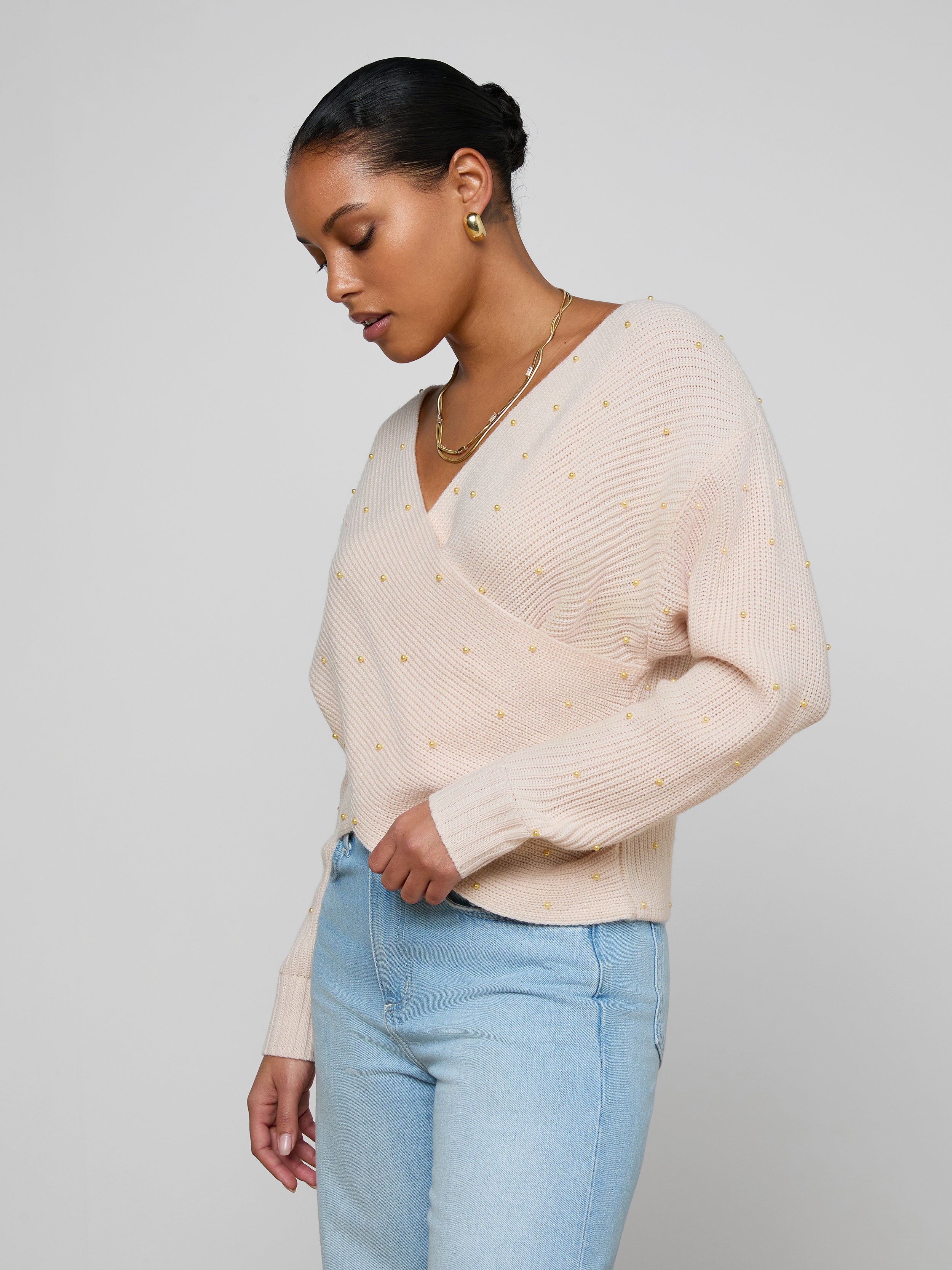 A woman wears the Antonella Crossover Sweater in light pink, featuring small textured dots, paired with light blue jeans. She stands against a plain off-white background, looking down and slightly to the side.