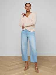 A woman with short hair poses confidently against a plain white background, one arm resting on her opposite elbow, wearing the Antonella Crossover Sweater – an oversized wool design – paired with light blue wide-leg jeans and beige heeled sandals.