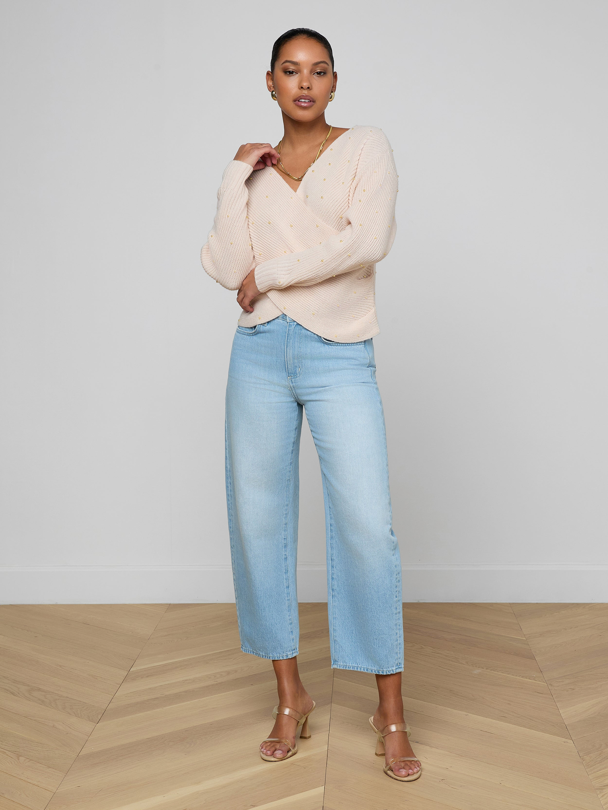 A woman with short hair poses confidently against a plain white background, one arm resting on her opposite elbow, wearing the Antonella Crossover Sweater – an oversized wool design – paired with light blue wide-leg jeans and beige heeled sandals.