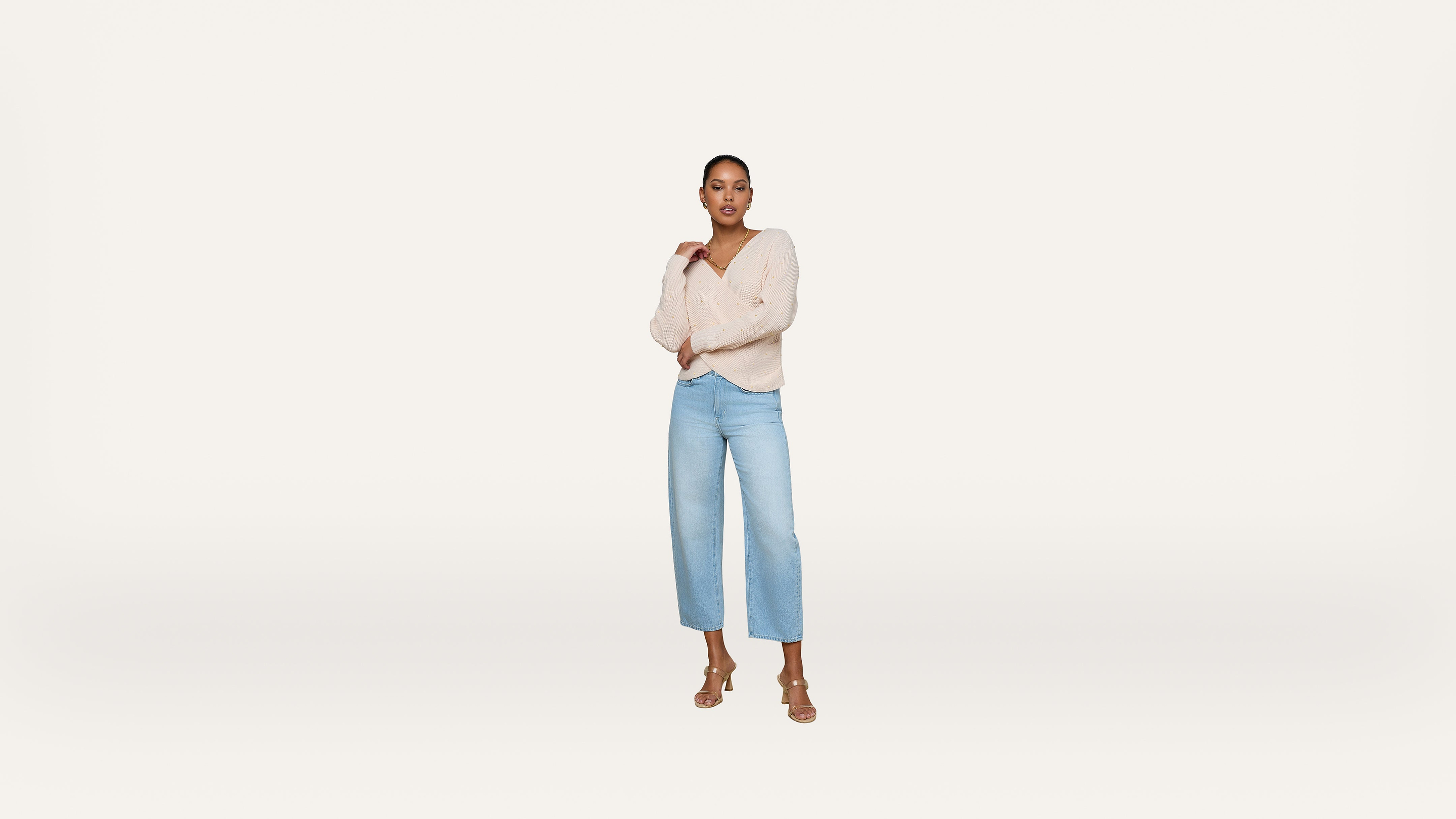 A woman stands confidently against a plain white background, wearing a light beige sweater, light blue high-waisted jeans, and tan heeled sandals. She poses with one hand on her chest and the other by her side.