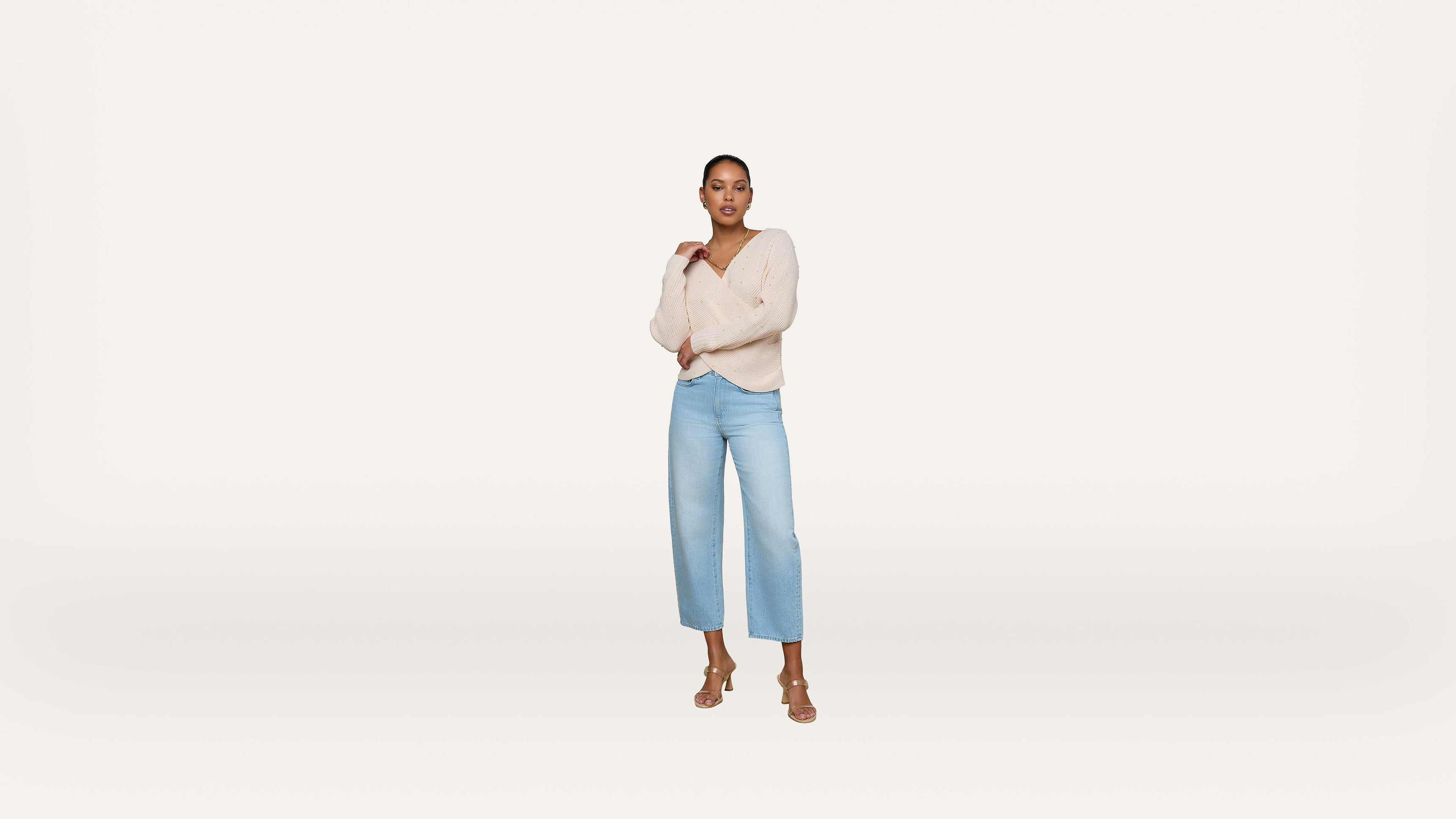 A woman stands confidently against a plain white background, wearing a light beige sweater, light blue high-waisted jeans, and tan heeled sandals. She poses with one hand on her chest and the other by her side.