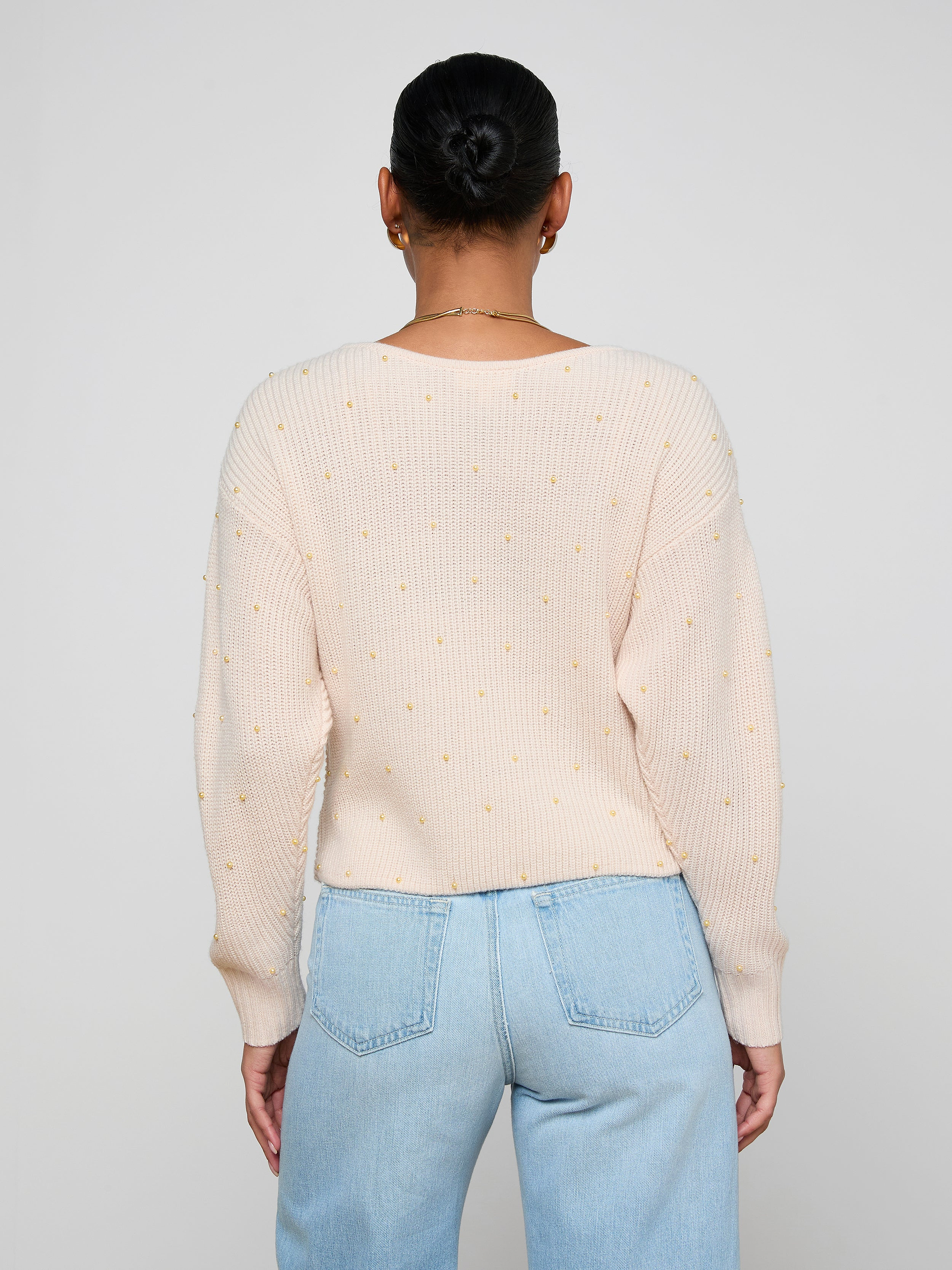 A person with dark hair in a neat bun is shown from behind, wearing the Antonella Crossover Sweater—a light pink knit featuring small yellow dots—paired with light blue jeans, standing against a plain off-white background.