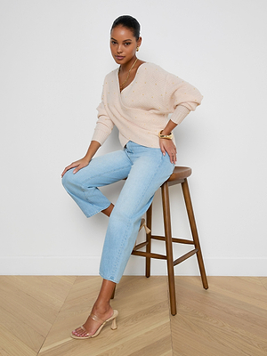 A woman sits on a wooden stool against a white wall, wearing the Antonella Crossover Sweater in light pink with gold dots, light blue jeans, gold jewelry, and clear-strap heeled sandals, looking at the camera in a relaxed pose.