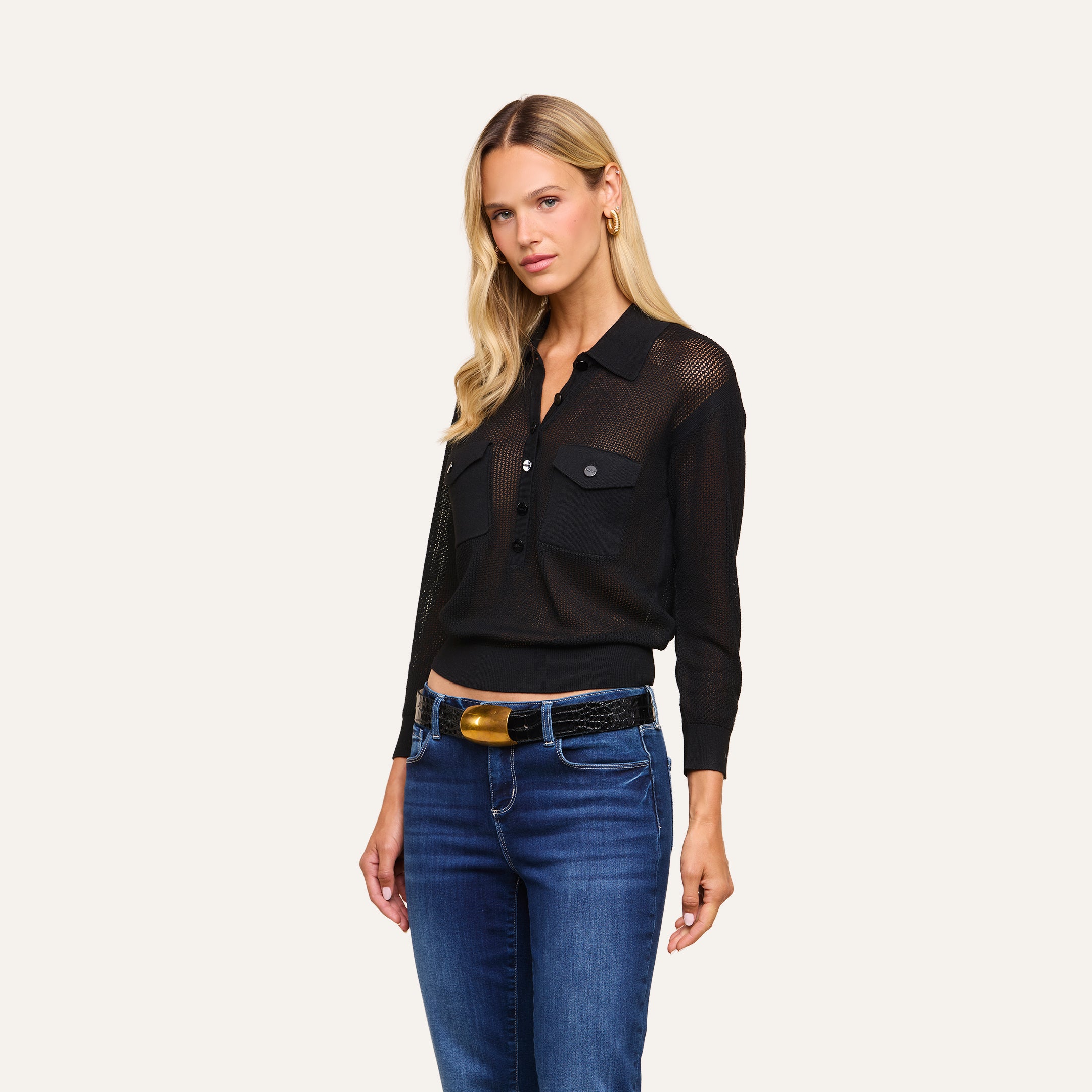 A woman with long blonde hair wears the black Carine Top, a lightweight knit design featuring a button placket, paired with blue jeans. She stands facing forward with a neutral expression against a plain light background.
