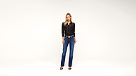 A woman stands against a plain white background, wearing a black long-sleeve shirt, blue jeans, black heels, and a black belt with a large gold buckle.