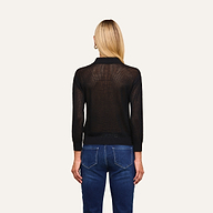 A person with long blonde hair, wearing the Carine Top—a lightweight black sheer knit long-sleeve top—paired with blue jeans, stands facing away from the camera against a plain white background.