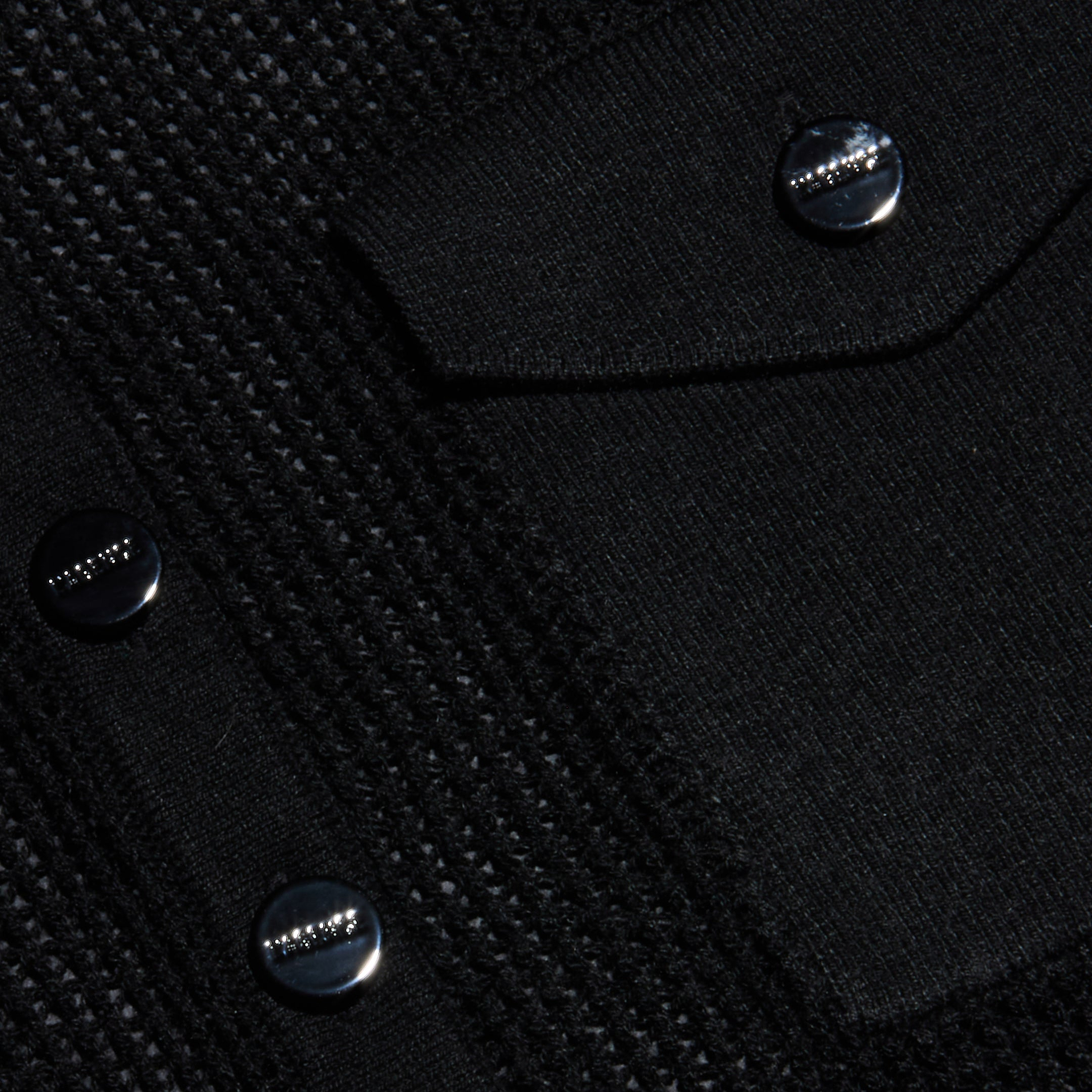 Close-up of the Carine Top’s textured black fabric, featuring shiny black buttons with a row of small raised metallic studs and a flap pocket. The surface combines lightweight knit textures with smooth patterns for added visual interest.