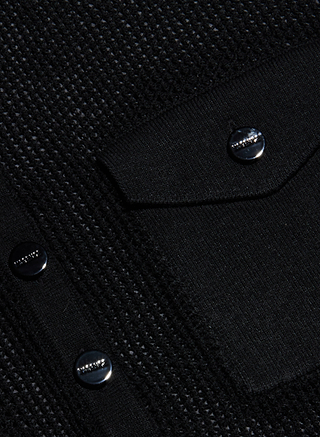 Close-up of a black textured fabric featuring a patch pocket with a flap. The pocket and flap are secured by shiny black buttons with a metallic accent, showcasing the refined details of this Carine Top in a lightweight knit.
