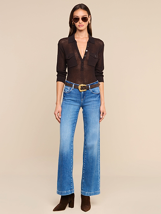 Woman wearing sunglasses, a sheer black long-sleeve Carine Top with front pockets layered over a lightweight knit sweater in coffee brown, blue high-waisted wide-leg jeans, a chunky belt, and brown heeled boots, standing against a plain beige background.