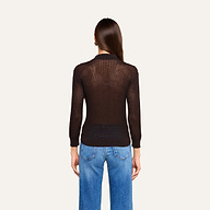 Une personne aux longs cheveux châtains, vêtue dun haut noir transparent à manches longues superposé au haut « Carine » de couleur café, et dun jean bleu, se tient de dos devant un fond beige.