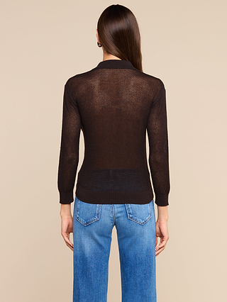 A person with long brown hair, wearing a sheer black long-sleeve top layered over the Carine Top in coffee brown and blue jeans, is standing facing away against a beige background.