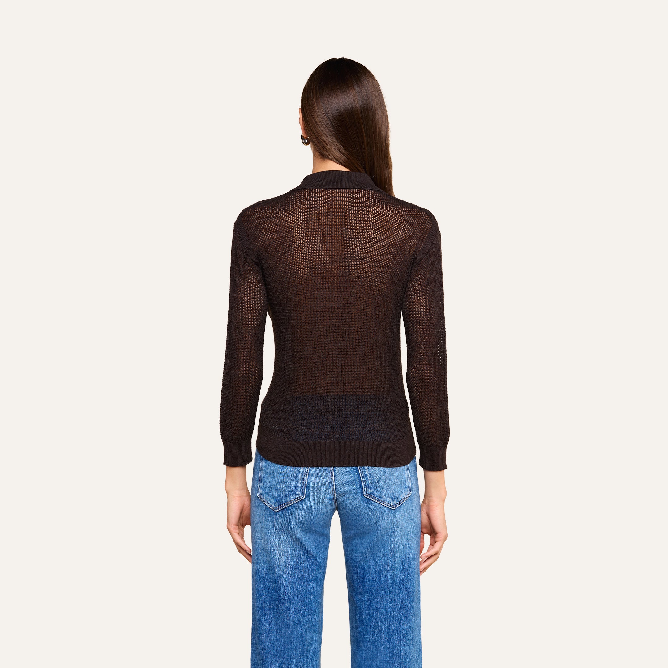 Une personne aux longs cheveux châtains, vêtue dun haut noir transparent à manches longues superposé au haut « Carine » de couleur café, et dun jean bleu, se tient de dos devant un fond beige.
