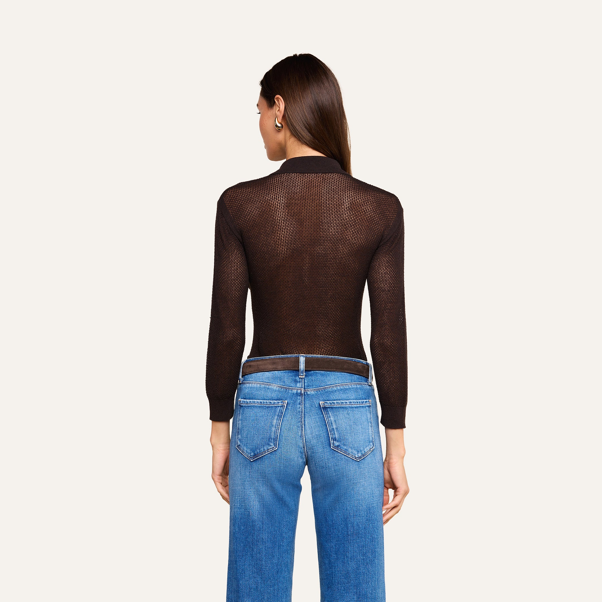 Une femme est de dos ; elle porte un haut « Carine » en maille légère, noir transparent et à manches longues, associé à un jean bleu. Larrière-plan est dun beige uni, et ses cheveux bruns, lisses, sont lâchés.