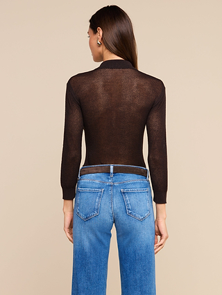 A woman stands facing away, wearing a lightweight knit Carine Top in sheer black with long sleeves, paired with blue jeans. The background is plain beige, and her straight brown hair is worn down.