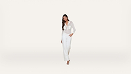 A woman with long dark hair stands against a plain white background, wearing a sheer white blouse, white pants, and open-toed heels. She has one hand in her pocket and is smiling slightly.