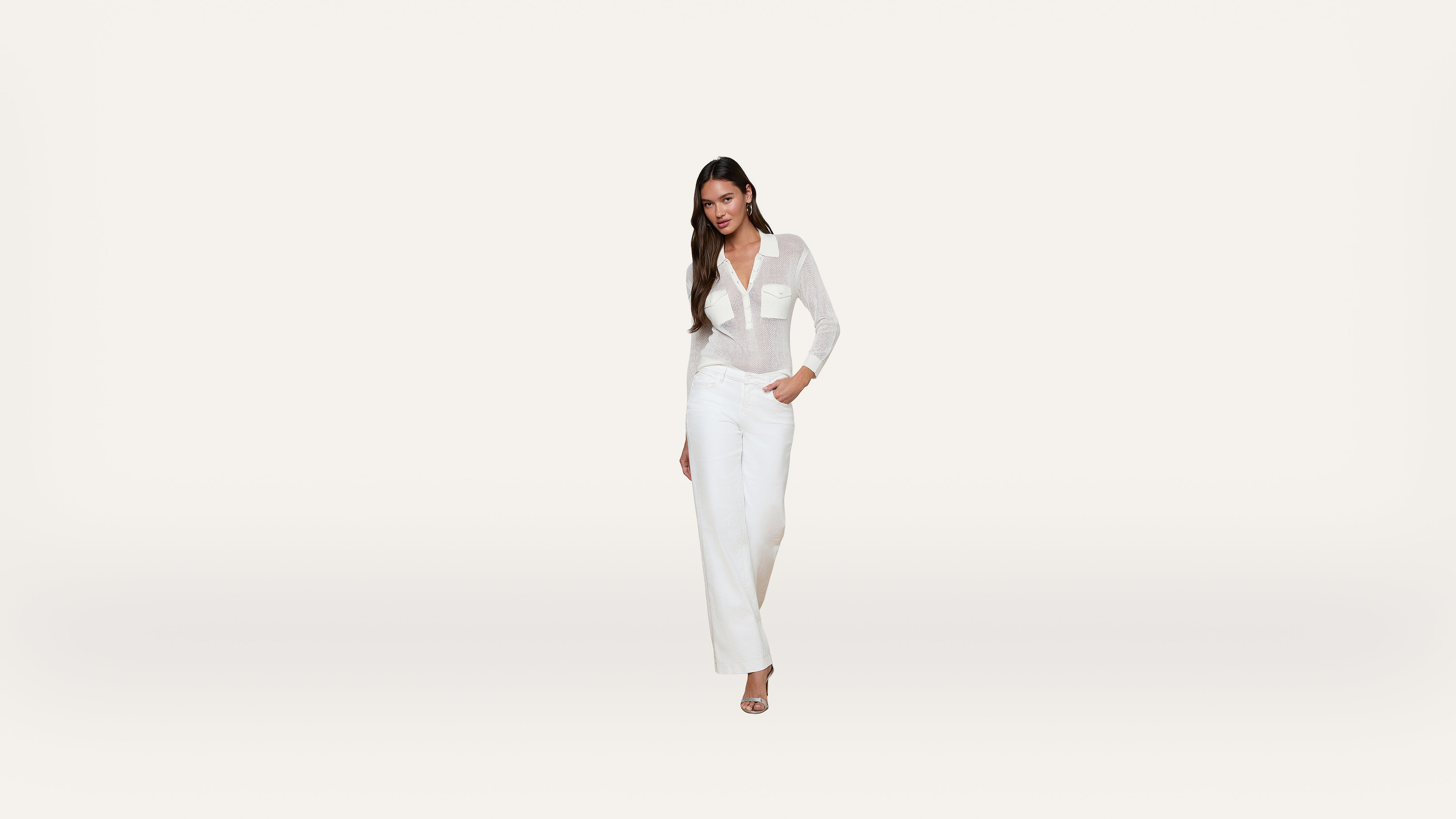 A woman with long dark hair stands against a plain white background, wearing a sheer white blouse, white pants, and open-toed heels. She has one hand in her pocket and is smiling slightly.