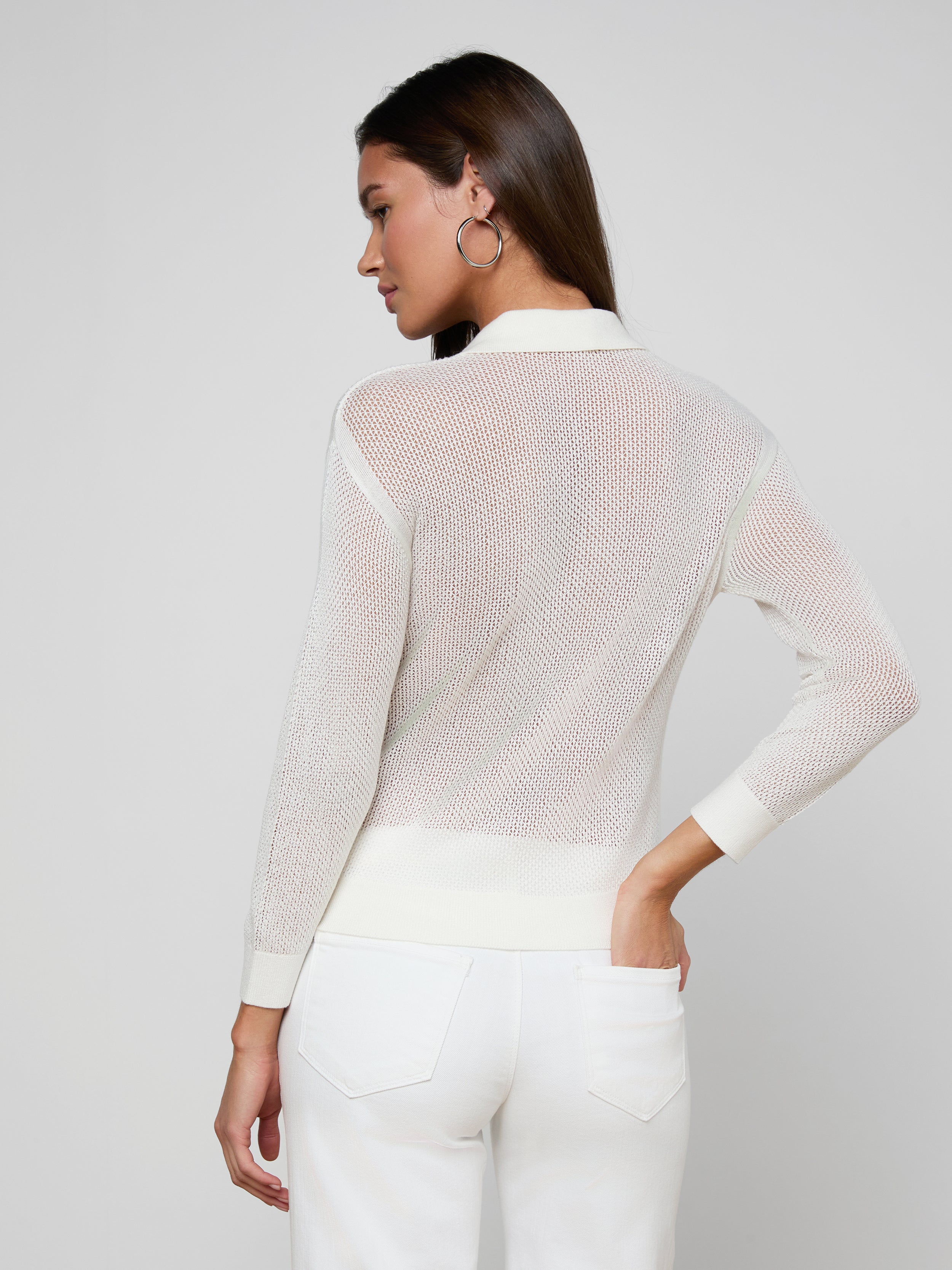 A woman stands with her back to the camera, wearing a sheer white long-sleeve Carine Top and white utility pocket jeans, one hand on her hip, against a plain light background.