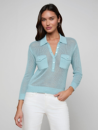 A woman with long dark hair wears a light blue Carine Top featuring a lightweight knit, button placket, and two chest pockets, paired with white pants. She stands against a plain light gray background.