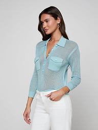A woman with long brown hair stands against a plain background, wearing a light blue Carine Top—a lightweight knit with front pockets, button placket, and white pants. She looks slightly to the side.