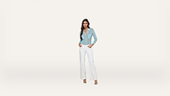 A woman stands against a plain white background, wearing a light blue, long-sleeve top and high-waisted white pants, with her hands in her pockets and looking forward.