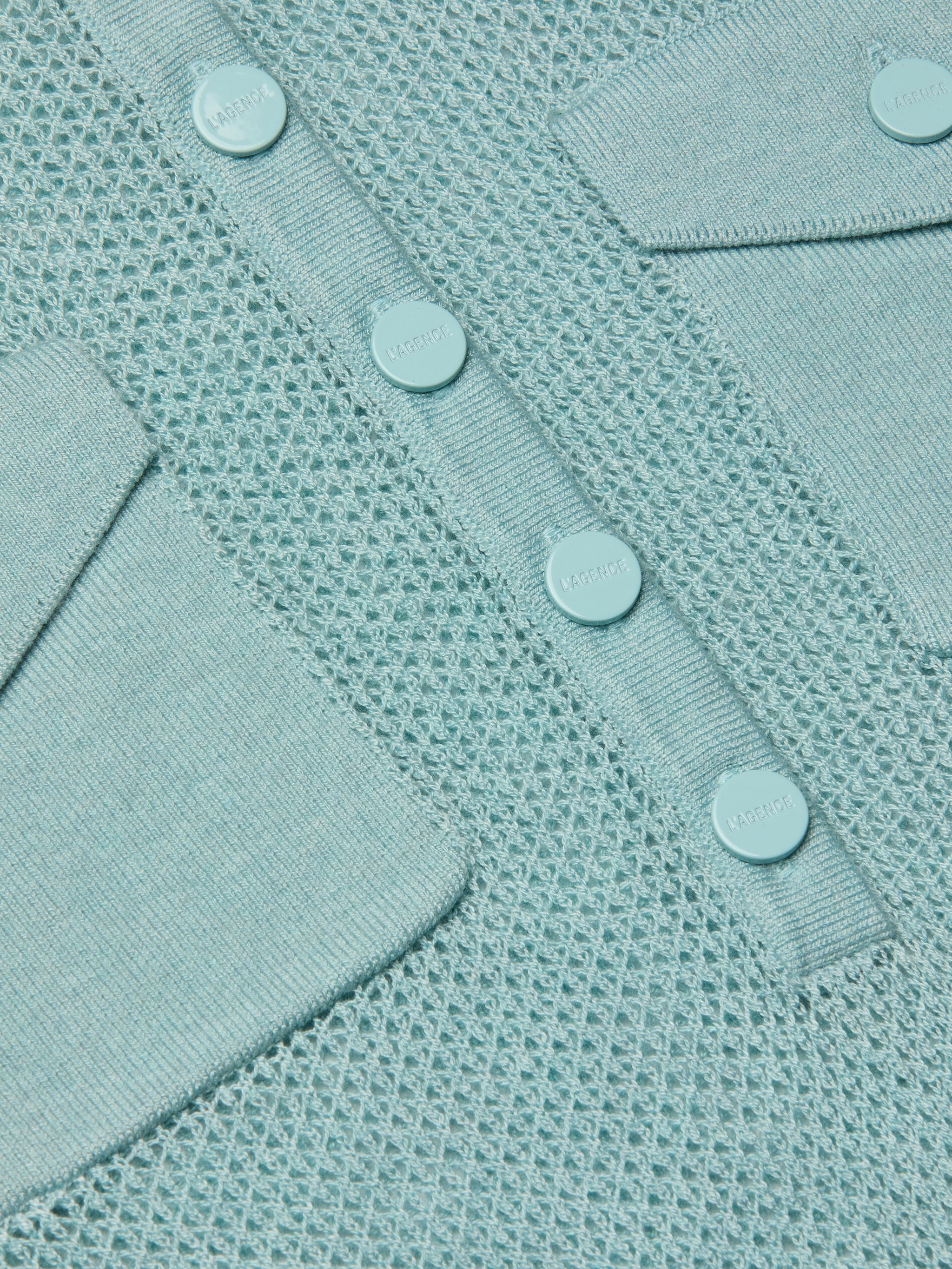 Close-up of the Carine Top in light blue, a lightweight knit with mesh pattern, button placket featuring four round buttons, and two angled flap pockets—all crafted from matching light blue material.