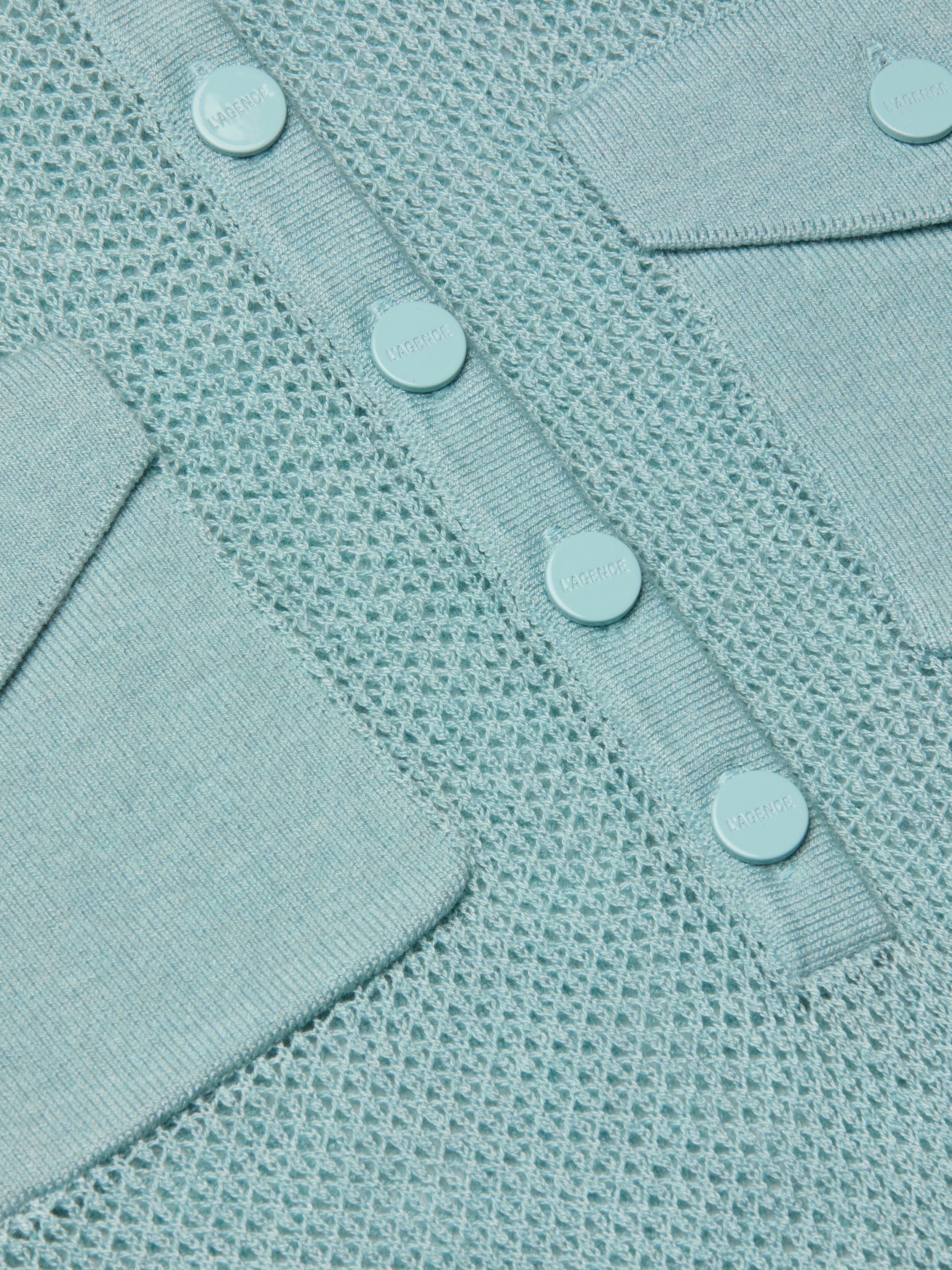 Close-up of the Carine Top in light blue, a lightweight knit with mesh pattern, button placket featuring four round buttons, and two angled flap pockets—all crafted from matching light blue material.
