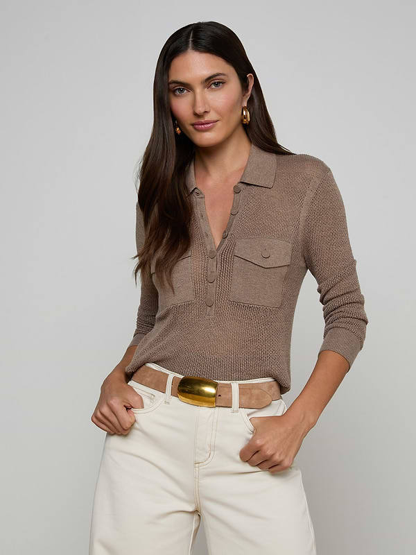A woman stands facing forward against a plain light background, wearing the Carine Top—a light brown, long-sleeve lightweight knit featuring utility pockets—paired with blue jeans. This everyday essential offers both style and practicality.