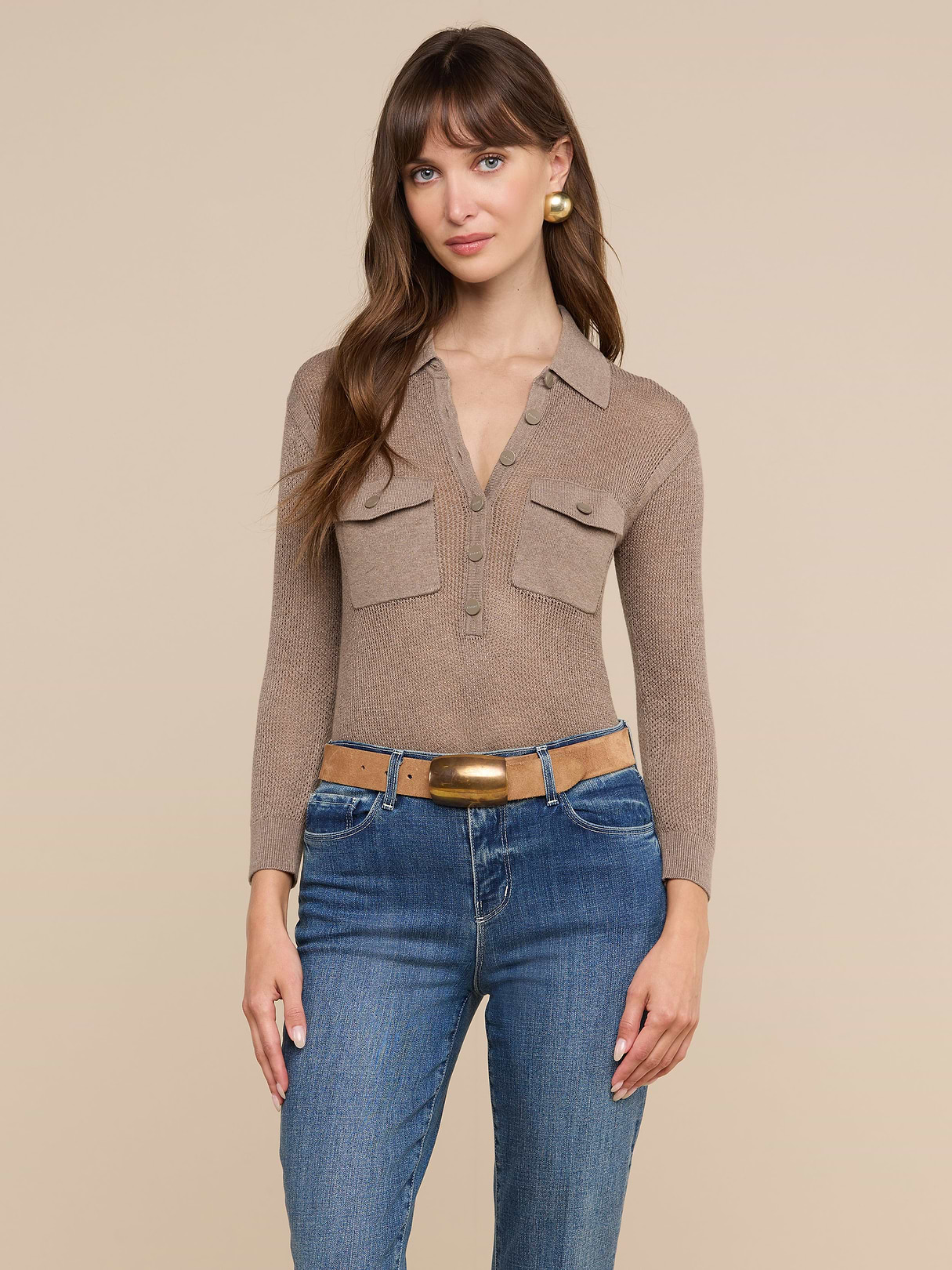 Une femme aux longs cheveux châtains porte le haut Carine — un modèle en maille légère couleur taupe doté de poches pratiques —, associé à un jean bleu, une ceinture marron et des boucles doreilles créoles dorées. Elle pose devant un fond beige pour un look décontracté de tous les jours.