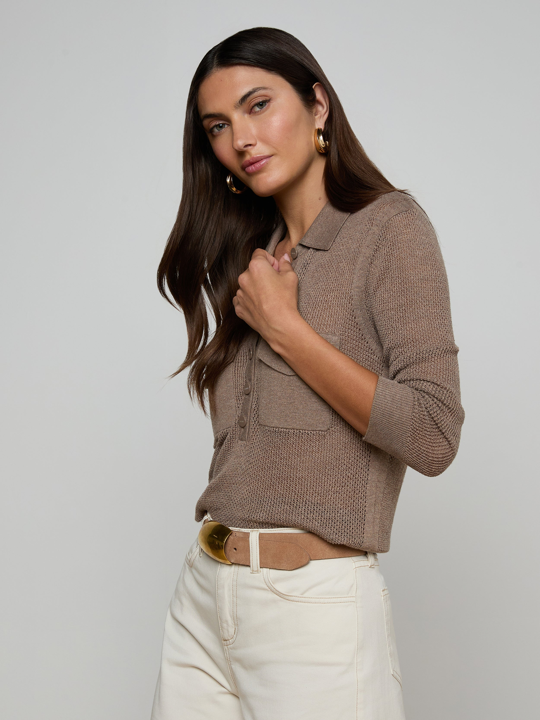 A woman poses sideways against a plain light background, wearing the Carine Top—a beige, long-sleeve lightweight knit with utility pockets—paired with blue jeans and one hand in her pocket.