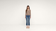 A woman with long brown hair stands facing forward, wearing a tan long-sleeve top, blue jeans, and tan heels, against a plain white background.