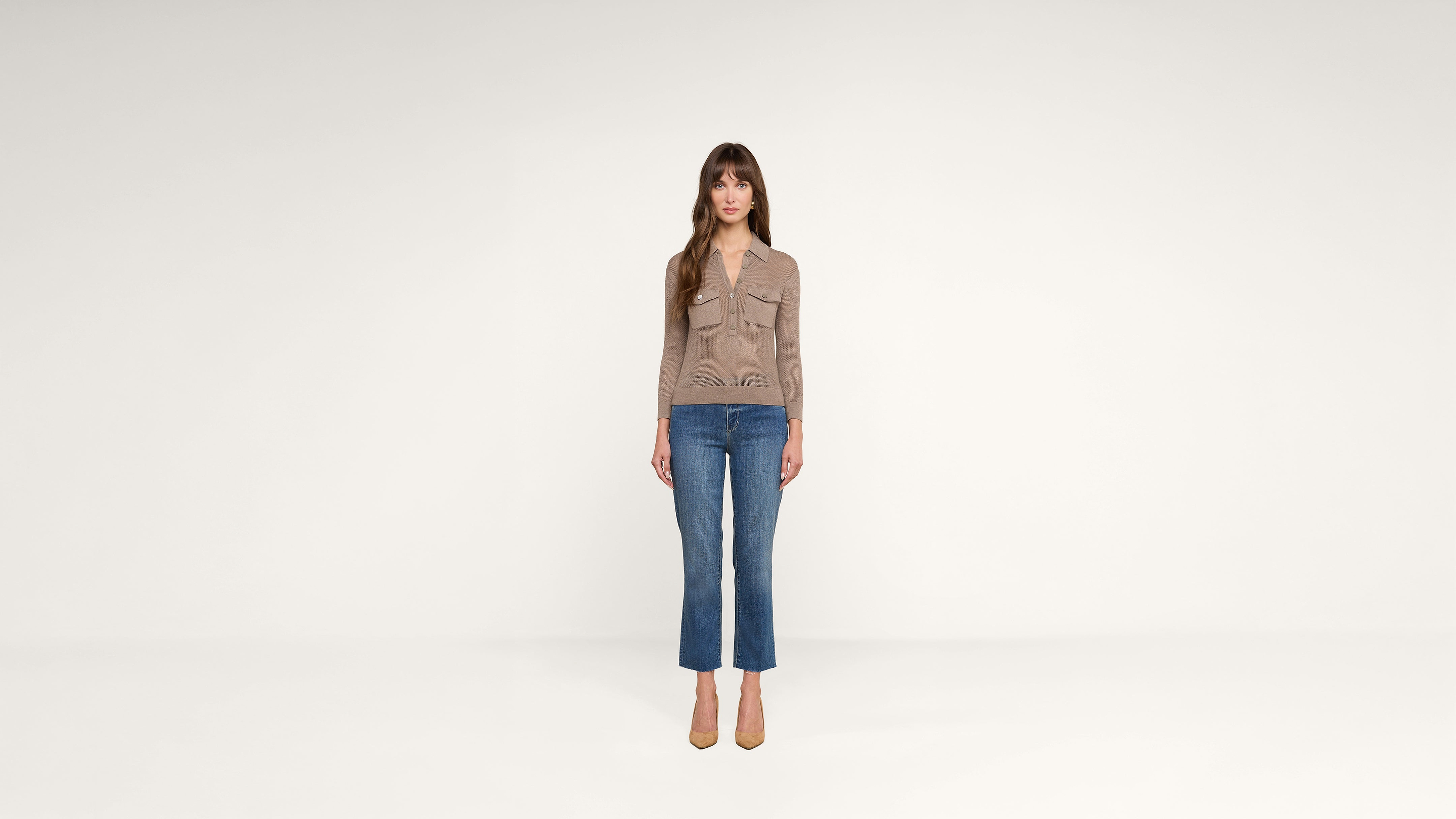 A woman with long brown hair stands facing forward, wearing a tan long-sleeve top, blue jeans, and tan heels, against a plain white background.