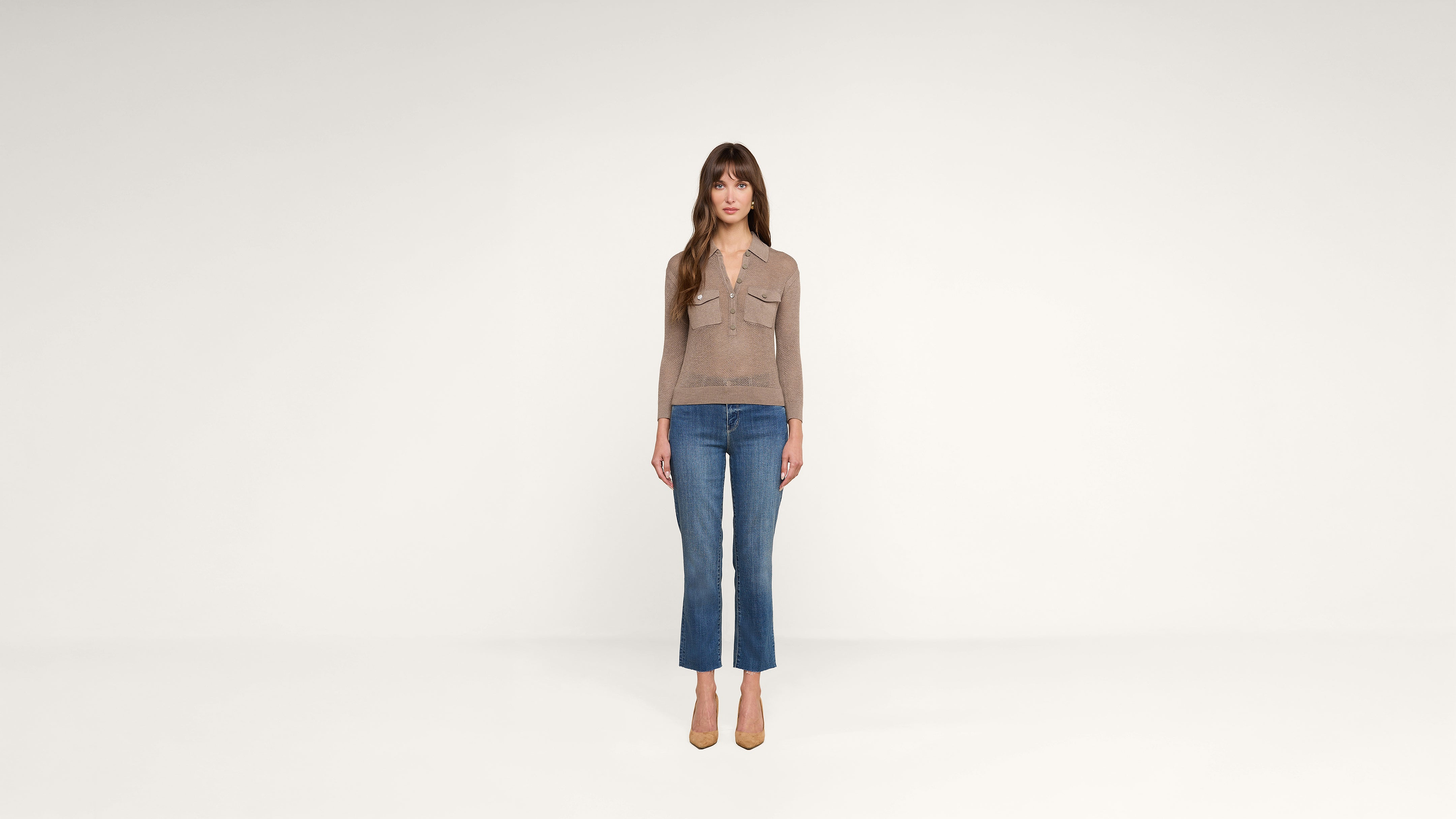 A woman with long brown hair stands facing forward, wearing a tan long-sleeve top, blue jeans, and tan heels, against a plain white background.