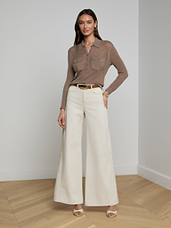 A woman wearing sunglasses, the Carine Top—a beige lightweight knit long-sleeve top—blue utility pocket jeans, a brown belt, and tan heels stands against a plain white background.