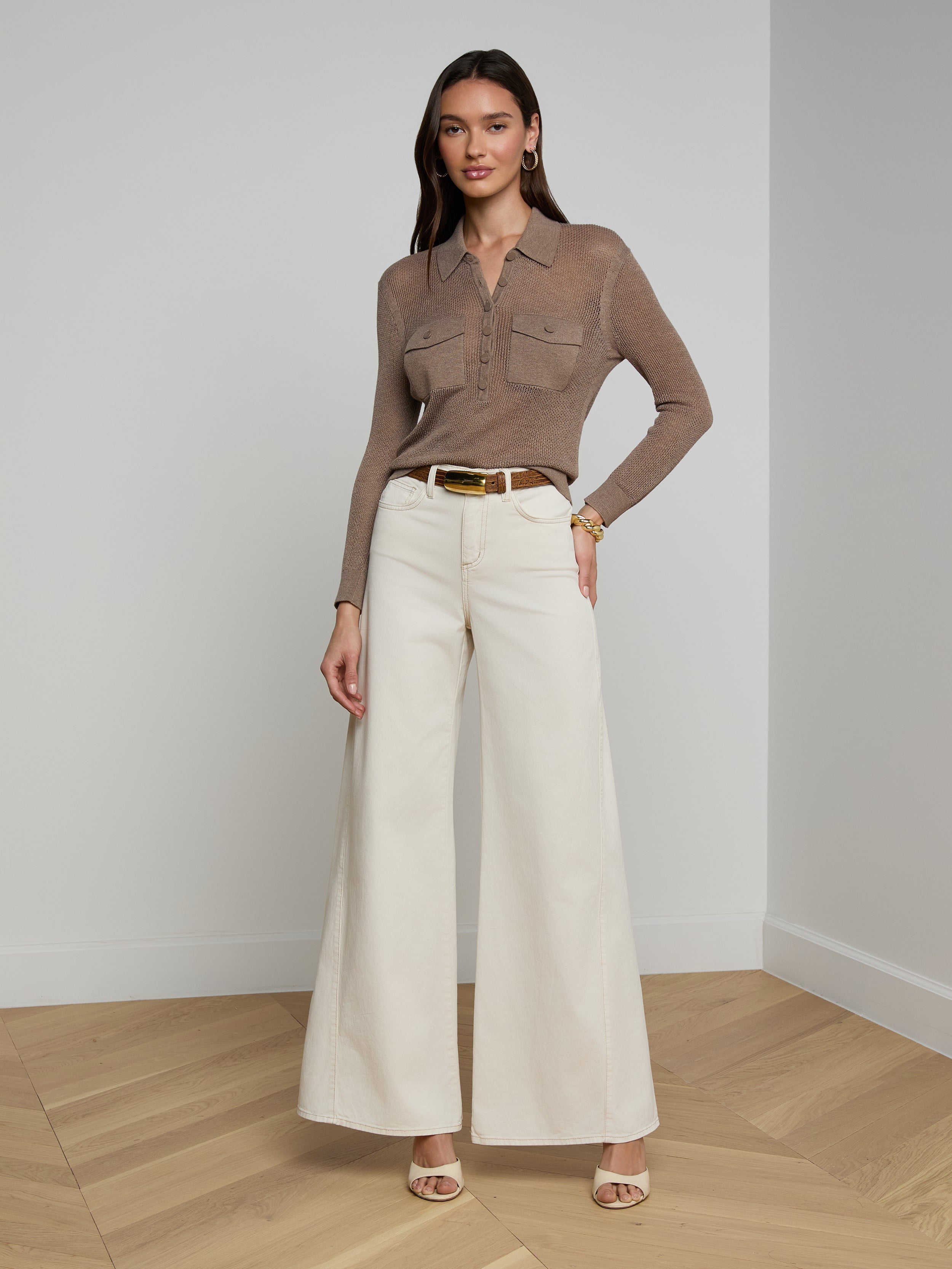 A woman wearing sunglasses, the Carine Top—a beige lightweight knit long-sleeve top—blue utility pocket jeans, a brown belt, and tan heels stands against a plain white background.