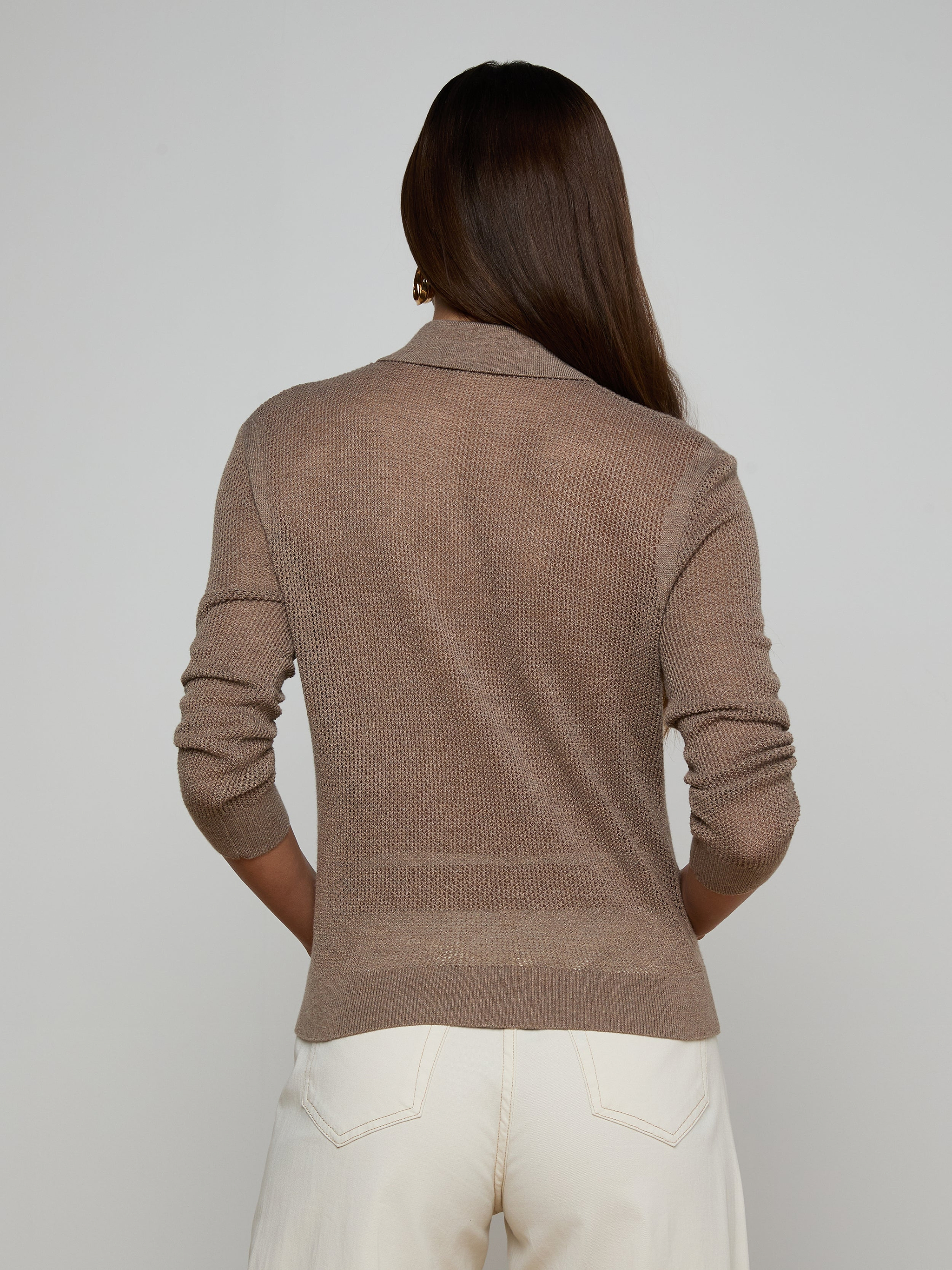 Wearing the Carine Top—a light brown lightweight knit long-sleeve sweater—a woman faces away, paired with blue utility-pocket jeans and standing against a plain light background.