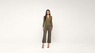 A woman stands against a plain white background, wearing a brown long-sleeve top, olive green cropped pants, and beige heels. She has long brown hair and is looking directly at the camera.