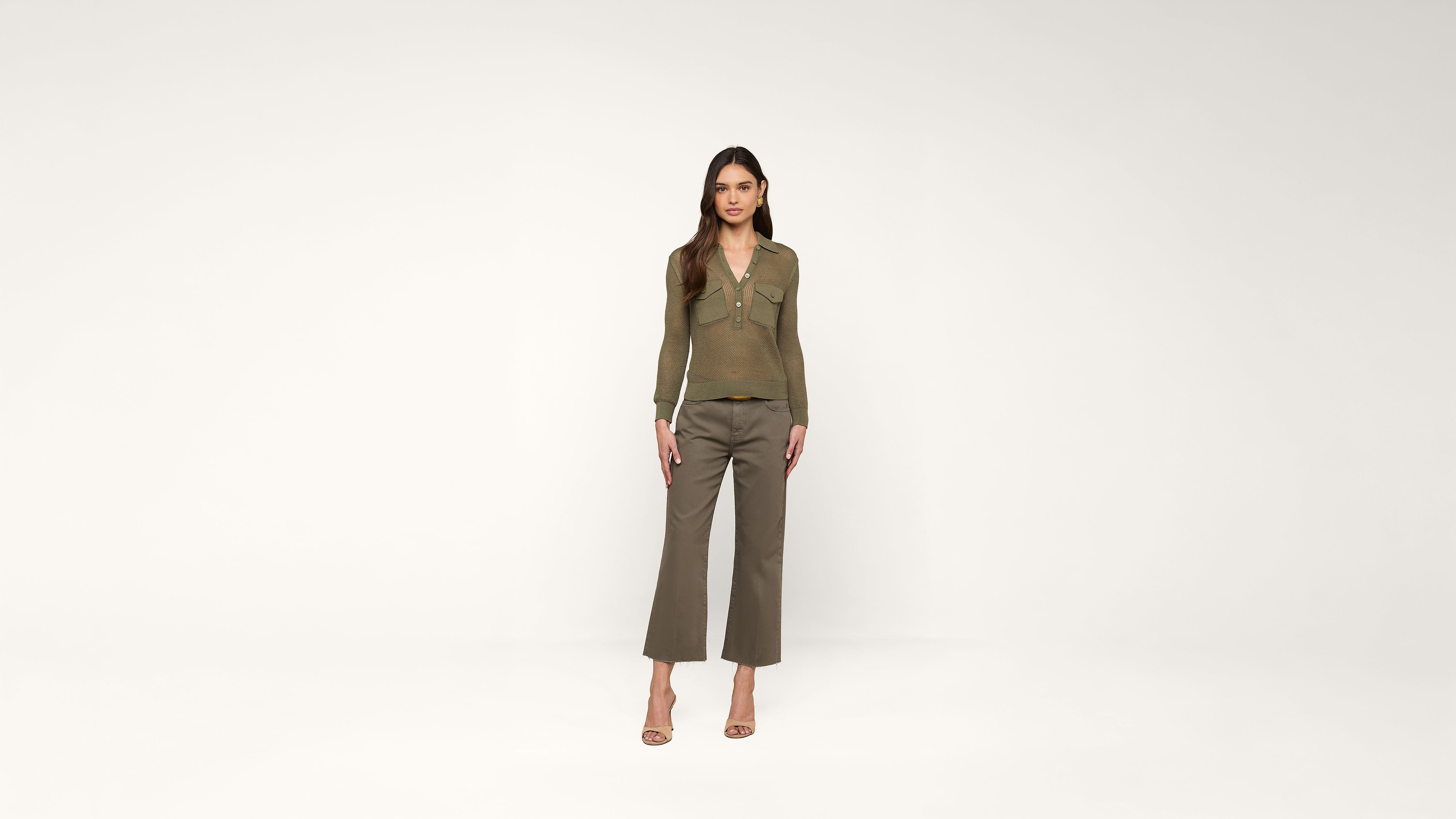 A woman stands against a plain white background, wearing a brown long-sleeve top, olive green cropped pants, and beige heels. She has long brown hair and is looking directly at the camera.