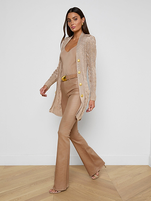 A woman stands indoors against a white wall and light wooden floor, wearing the Penelope Cabled Cardigan with gold logo buttons, paired with a tan top, tan flared pants, a wide belt, and heeled sandals.
