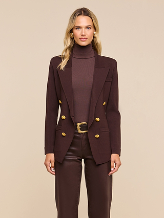 A woman stands facing forward against a beige background, wearing the Amelia Knit Blazer in dark brown with gold buttons and removable shoulder pads, paired with a matching turtleneck and leather pants.
