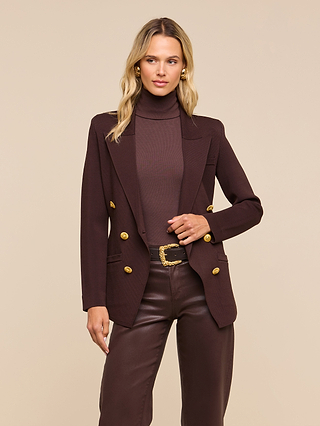 A woman wearing the Amelia Knit Blazer—a dark brown structured knit blazer with removable shoulder pads and gold buttons—along with a brown turtleneck and brown leather pants, stands against a beige background.