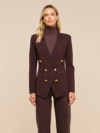 A woman stands against a beige background, wearing the Amelia Knit Blazer—a dark brown structured knit blazer with gold buttons and removable shoulder pads—layered over a matching turtleneck and pants. She accessorizes her loose blonde hair with gold earrings.