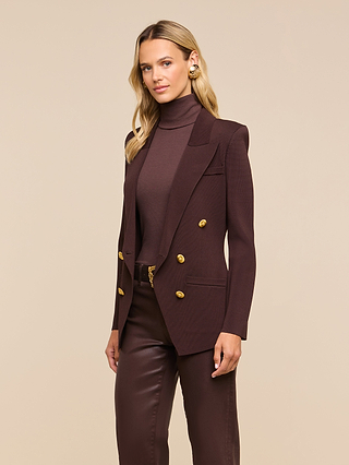 A woman stands against a beige background, looking at the camera with a slight smile, wearing the Amelia Knit Blazer in dark brown—featuring gold buttons and removable shoulder pads—paired with a matching turtleneck and leather pants.