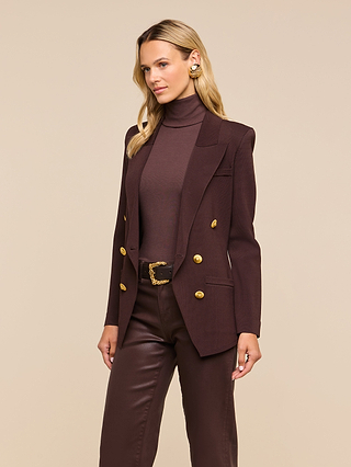 A woman wearing the Amelia Knit Blazer—a dark brown, structured knit blazer with removable shoulder pads and gold buttons—paired with a matching turtleneck and brown pants stands against a beige background.