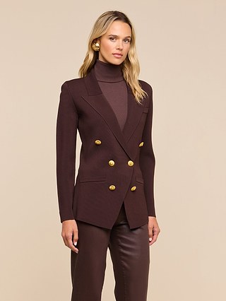 A woman stands against a beige background wearing the Amelia Knit Blazer—a dark brown, structured knit blazer with gold buttons and removable shoulder pads—layered over a matching turtleneck sweater and paired with brown pants.