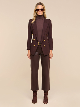 Woman wearing sunglasses, the Amelia Knit Blazer with removable shoulder pads and gold buttons, a matching dark brown turtleneck, dark brown pants, and brown suede boots, standing against a plain beige background.