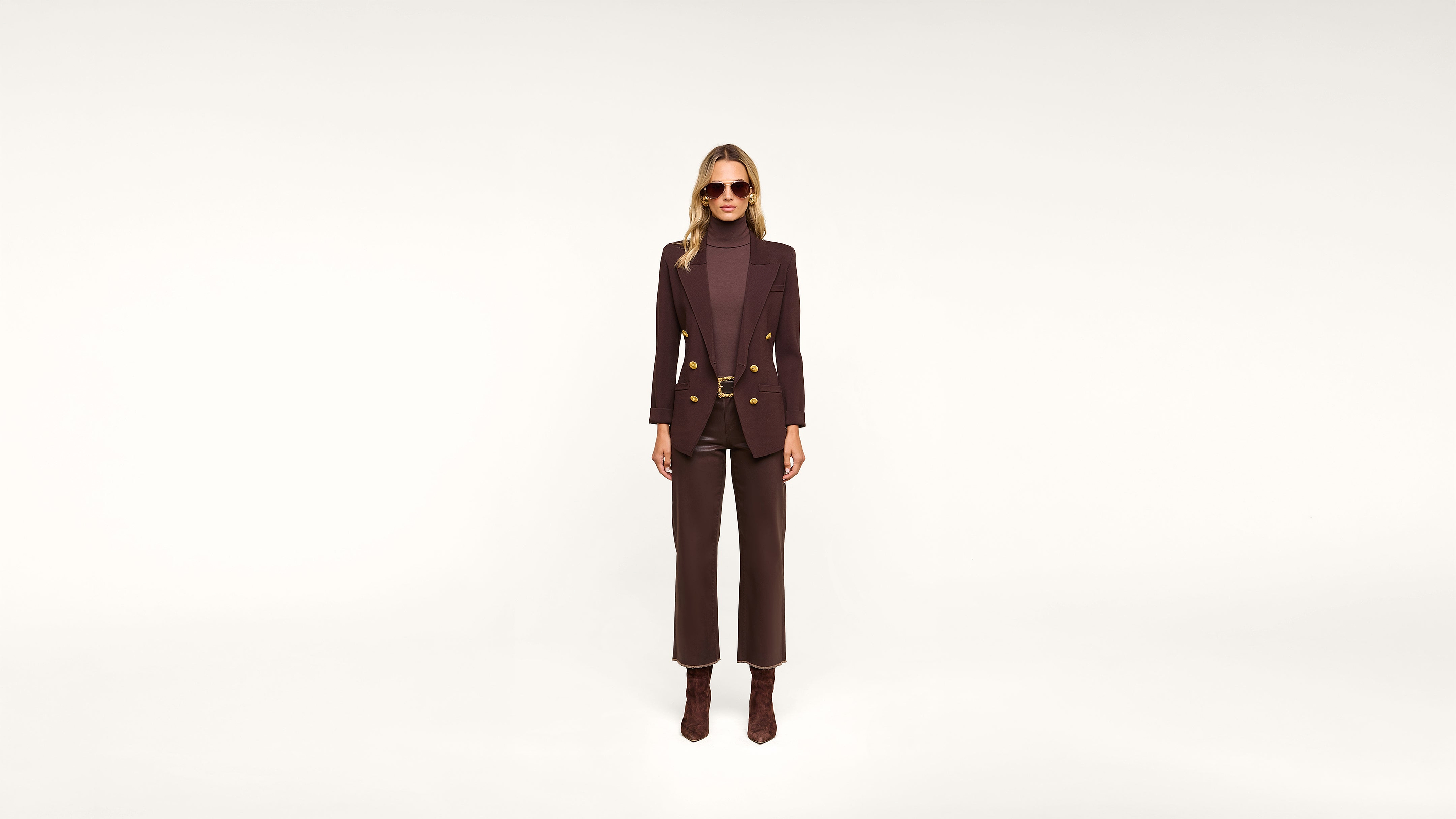 A person wearing sunglasses, a dark blazer with gold buttons, matching pants, a turtleneck, and boots stands against a white background.