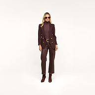 A woman wearing sunglasses, the Amelia Knit Blazer in dark brown with gold buttons crafted in soft tailoring, a matching turtleneck, brown pants, and ankle boots stands against a plain white background.