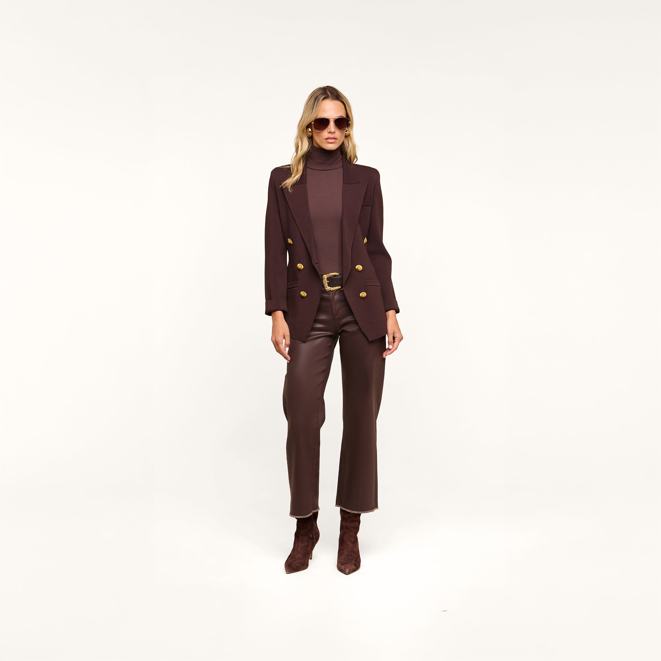A woman wearing sunglasses, the Amelia Knit Blazer in dark brown with gold buttons crafted in soft tailoring, a matching turtleneck, brown pants, and ankle boots stands against a plain white background.